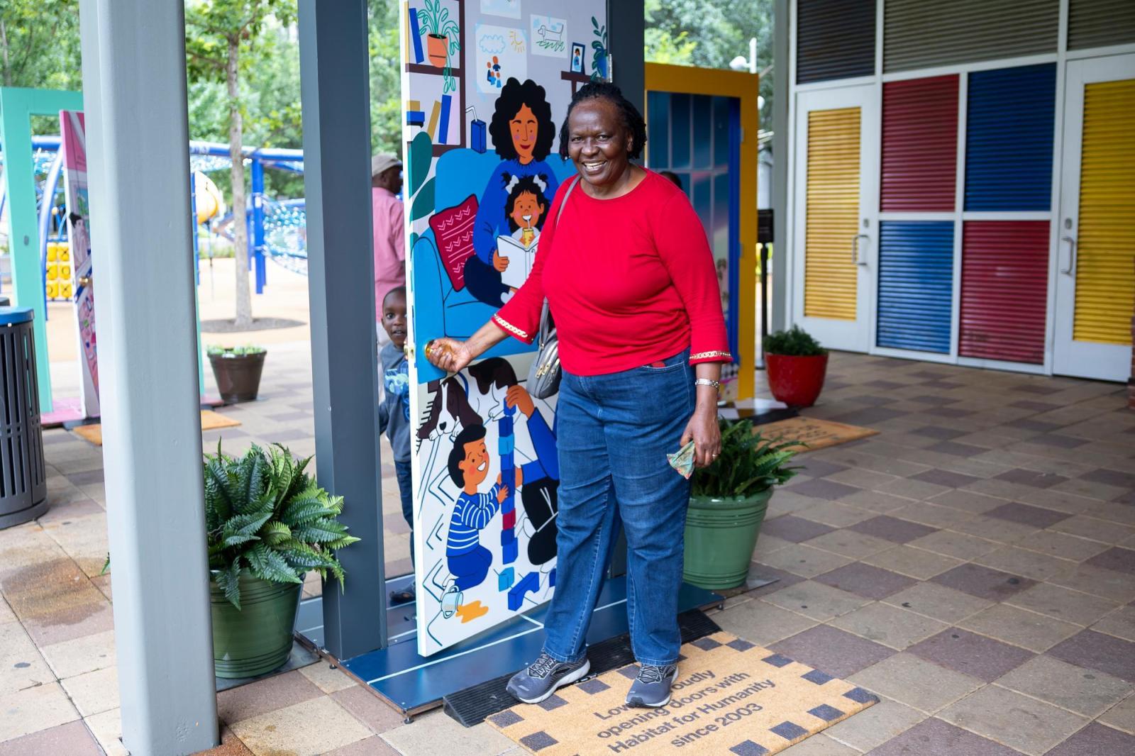 An older woman in a red shirt and jeans posing with an illustrated door while a little boy peaks through the doorway. 