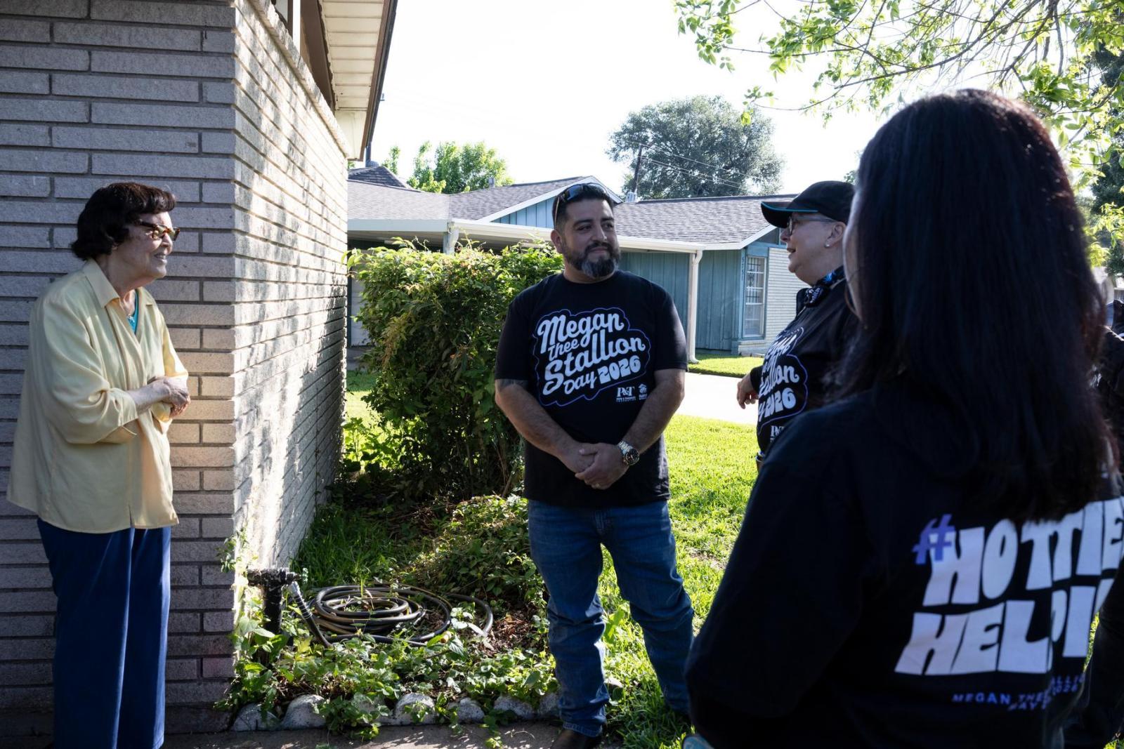 Volunteers in black shirts stand in front of a white brick home talking to the homeowner.