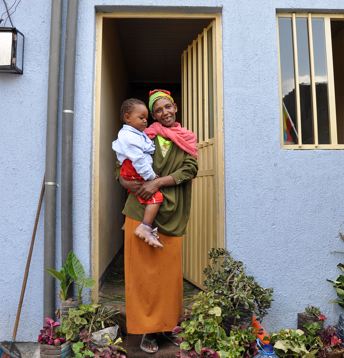 A woman, Yiftu, stands in the doorway of her home, holding a child and smiling. They are surrounded by planters.
