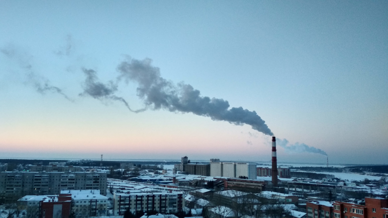 A snowy city skyline with a factory smokestack rising high above the buildings emitting smoke