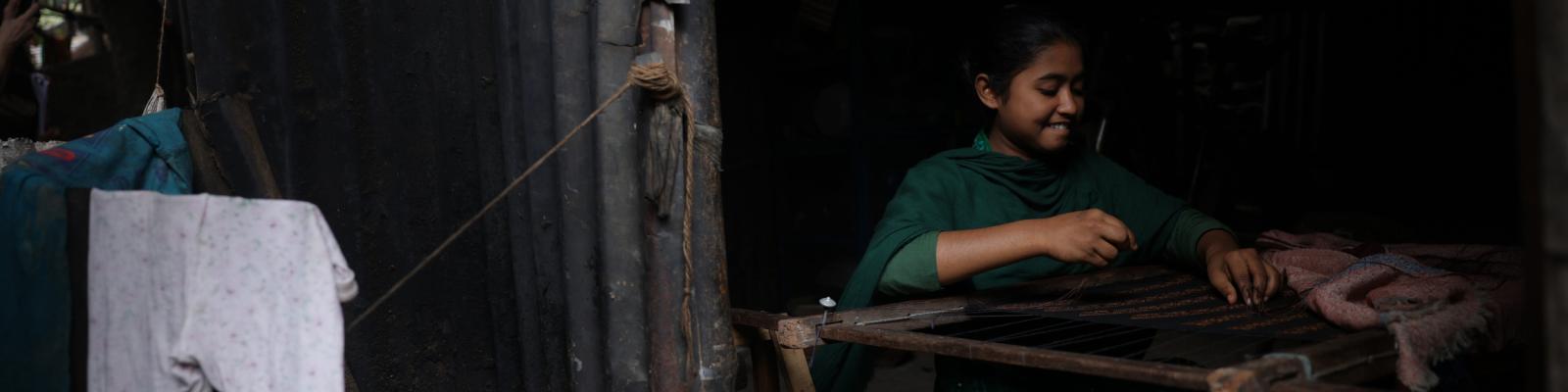 Young girl in green clothing smiling as she weaves on a loom inside her home