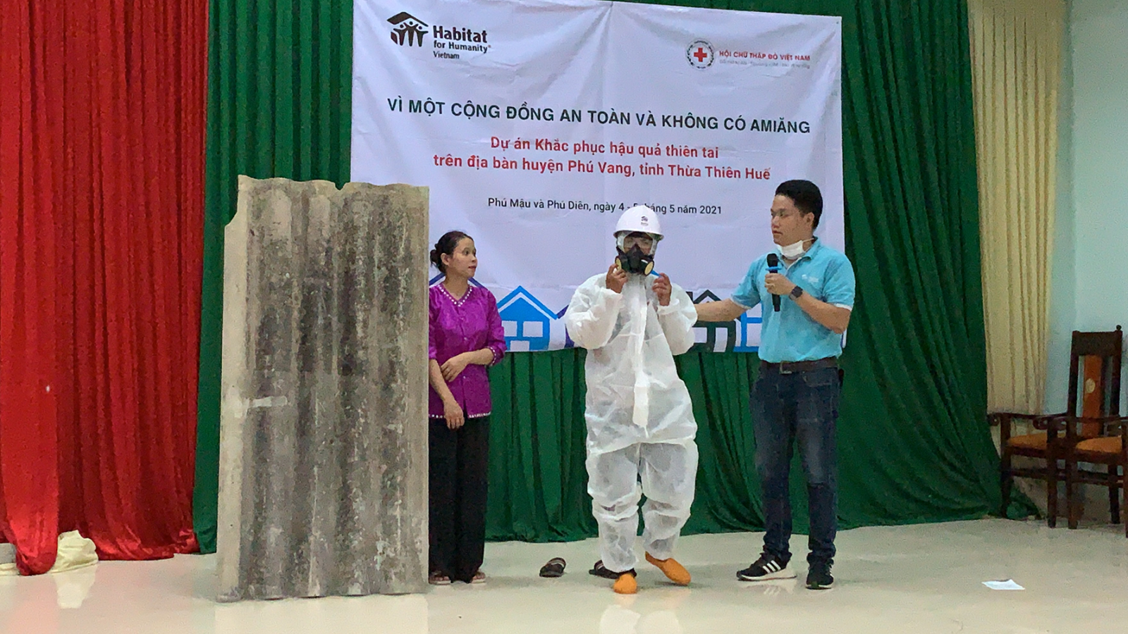 Three people doing a presentation about asbestos safety. One is wearing a white hazmat suit. 