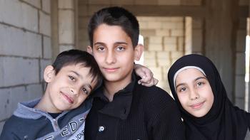 siblings from Syria living in Lebanon - two boys and a smiling girl wearing black scarf, new brick wall in the background