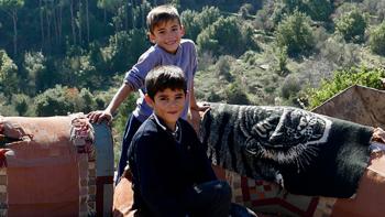 Photo: two boys sitting on an old sofa outside. Mountains in the background.