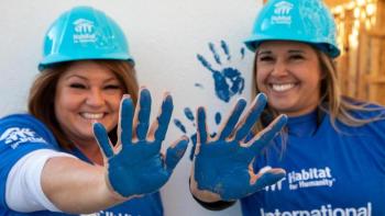 Two smiling women volunteers in hardhats hold up their hands which are painted blue.