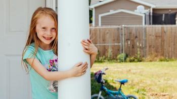 Young girl with red hair smiles and hugs a column on front of her Habitat house.