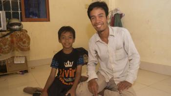 A man and child kneel on the floor of their home together, smiling.