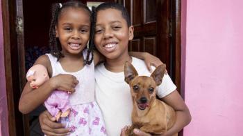 Young girl holding doll smiles with her brother in their doorway holding small dog.