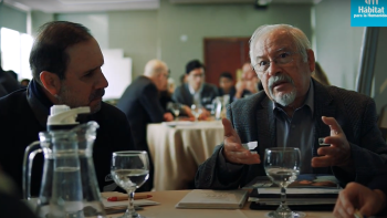A man talks to a group while sitting at a table.