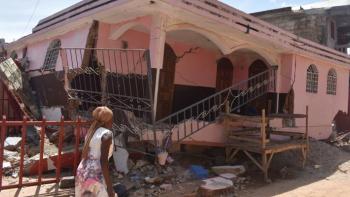 A woman walks by a partially collapsed house in Haiti.