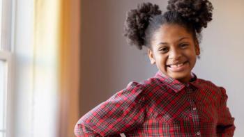 A young girl with pigtails stands by a window smiling.