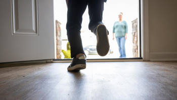 A child, visible from the legs down, runs through the open door of their home to their mother waiting outside.