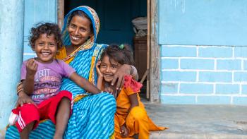 Woman in blue and ochre sari sits with her children on the porch of their home. The children are smiling and laughing with their mother. In the background is a light blue wall and an open doorway.