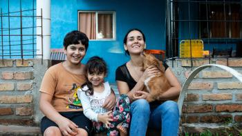 Woman sitting with her two children and holding a small dog. The family is smiling and they are framed by the vibrant blue wall of the house.