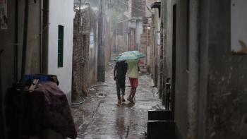Two people sharing an umbrella in heavy rain walk away from the camera down an alley in Duaripara, an informal settlement in Dhaka, Bangladesh.
