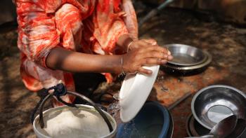 Hands of person in orange garment crouched down and washing dishes at the site of the community well.
