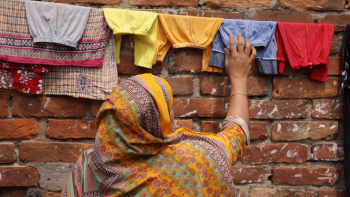 Woman in yellow-orange patterned hijab reaching up toward a clothesline where colorful garments are drying. Behind the clothesline is a brick wall. 
