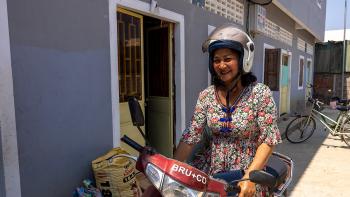 A smiling woman in a floral dress and a helmet prepares to ride her motorcycle in front of her home in Cambodia.