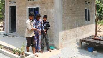A man and boy on either side of an elderly woman, helping her walk down a ramp walkway out of her house.