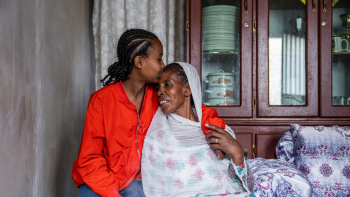 A woman sits in a white dress with a scarf over her head and her daughter sits beside her kissing her head.