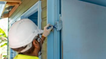 A volunteer in a white hard hat and yellow vest paints a front door with blue paint.