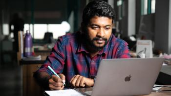 A man sitting at a desk in front of his laptop, writing on a notepad.