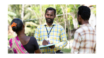 A man with a clipboard chats with two colleagues outside.