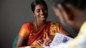 A woman in a sari sits at a desk smiling as a man in front of her reviews paperwork.