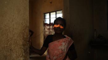 A woman stands in shadows with a swatch of light from the sun streaming through the window lighting up her eyes.