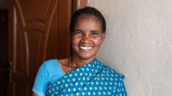 Devi, residente de Tamil Nadu, que ahora disfruta de un techo fresco, está de pie frente a una puerta y una pared blanca, vestida con un sari azul y sonriendo.
