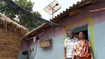 Couple standing in front of their colorful house, with a solar panel visible on the roof.