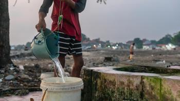 A person in a red shirt and black and white striped shorts draws water from a communal well with a broken blue bucket. 