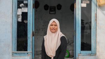 Woman in light tan hijab and black dress standing in the doorway of her rental home. The doorway and sidelights on either side of her are painted in a pale blue and the house is white.. 