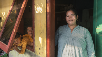A mother stands in an open front door while her children lean out of a window, all staring at the camera.