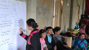 Woman in red scarf writing on a large piece of paper on the wall. Several other people are seated behind her.