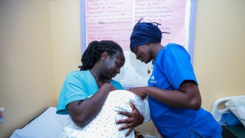Kenyan nurse dressed in blue scrubs checking on a Kenyan mother and her new baby in a clinic.