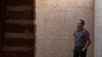 A man stands next to a brick wall of his newly built home with a look of pride on his face.