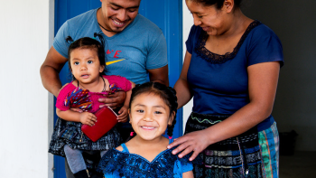 Family standing outside their white house with a bright blue door. A little girl is front and center smiling widely.