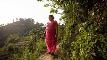Niru walks a path near her house. She is surrounded by green trees. She is wearing a pink sari and looking off into the distance.