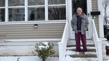 A man with gray hair standing on the porch steps in front of his beige home. 