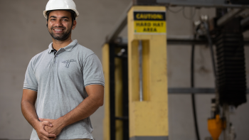 Sales and marketing executive Tushar Joshi stands in front of the TVASTA offsite 3-D production printer. He is wearing a long sleeve button down shirt and a white hard hat.