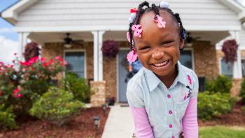 A little girl laughing outside her home with a lush green yard.