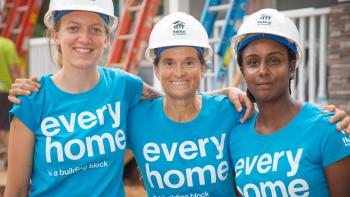 Three volunteers in blue shirts and hard hats smiling on a build site.