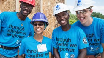 A group of four young people in blue shirts smiling on a build site.
