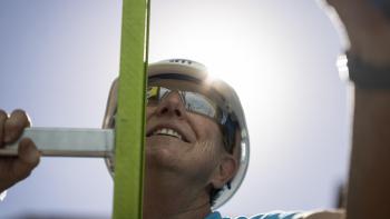 A Habitat volunteer in a blue shirt, white hard hat, and sunglasses lifts his arm above his head as he holds on to a yellow ladder.