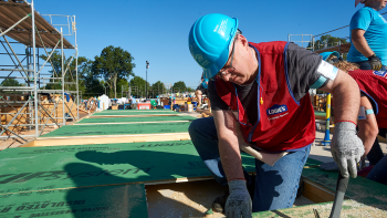 A man in a red Lowe's vest and a blue Habitat hard hat kneels down and hits a hammer into a piece of wood.