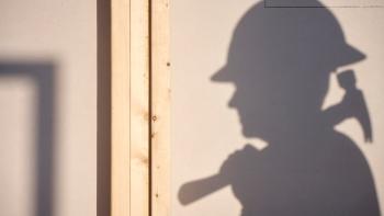 The shadow of a volunteer wearing a hard hat and holding a hammer appears on a white wall.