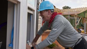 Jonathan Reckford wearing a blue hard hat on a Habitat build site, smiling as he leans in to an open window 