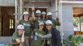 Group of young volunteers in army green Habitat for Humanity shirts and white hard hats in front of build site. 