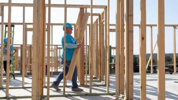 A volunteer in a blue Habitat shirt and hard hat carries an armload of lumber through the frame of a house.
